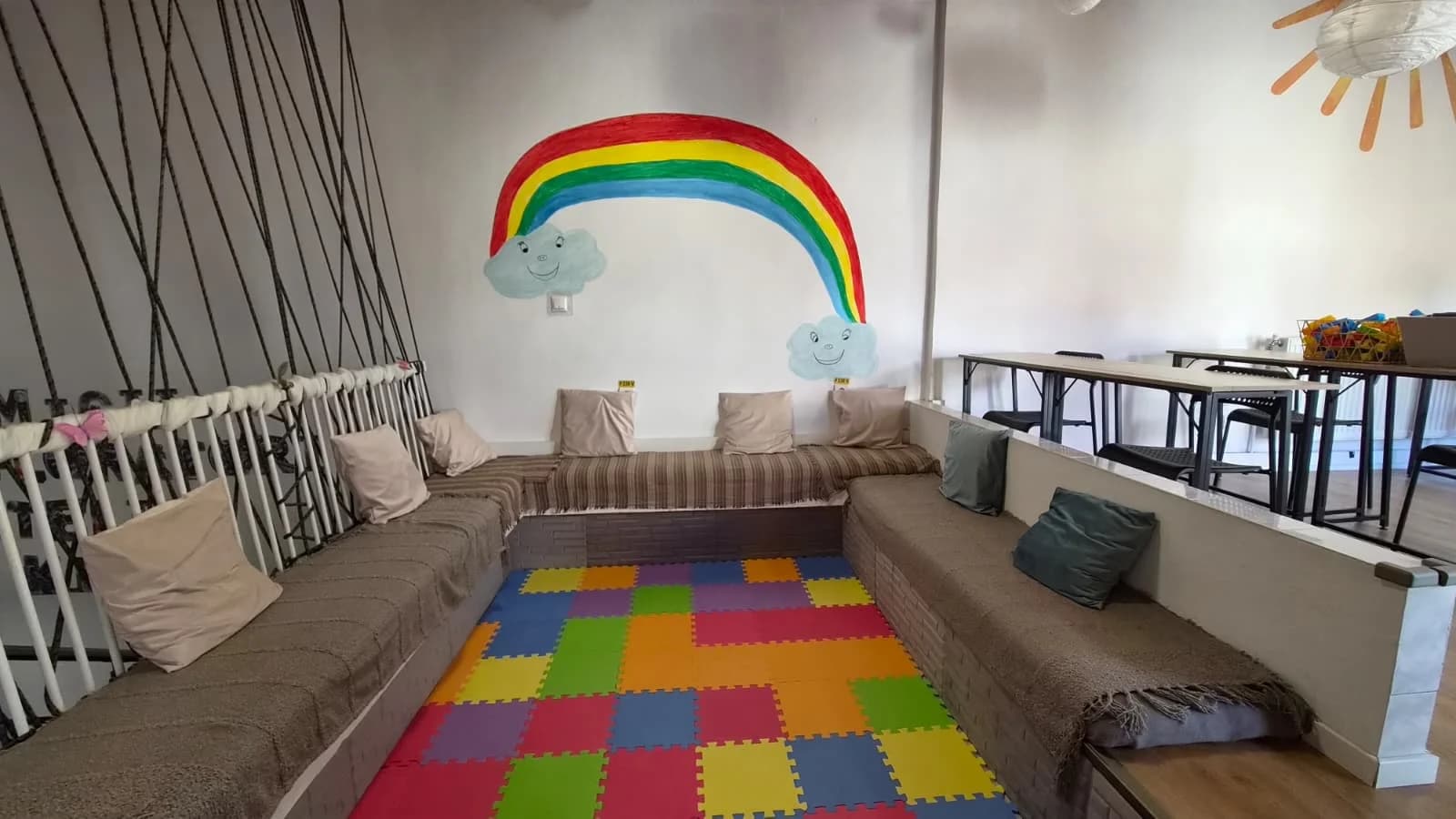 Colțul de joacă, cu pictura cu curcubeu pe perete, canapea cu perne și covor puzzle colorat